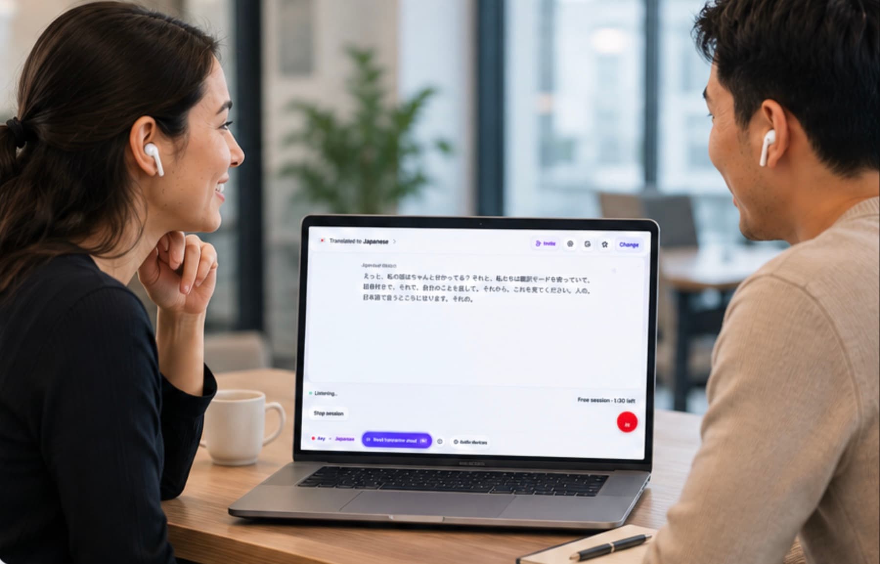 Two people talking across an office table with a laptop between them, showing their conversation translated into Japanese while each wears their own earbuds.